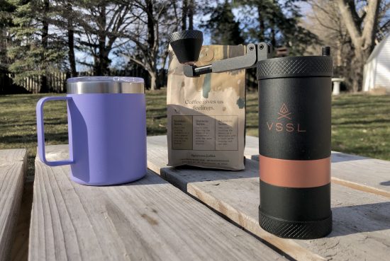 A closeup of a purple coffee mug, the middle a coffee bag, and to the right the hand grinder. They are all on a picnic table outside.
