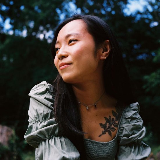 Sierra is a young woman with long black hair. Featured is a closeup portrait of her. She looks to the left smiling, and wears a puffy green blouse and a necklace. She has a tattoo of a coffee plant on her chest.