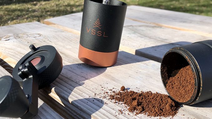 The VSSL hand grinder sits split apart on a picnic table outdoors. To the right is the already ground coffee in the chamber. In the middle is the grinding portion of the grinder and to the left is the handle.