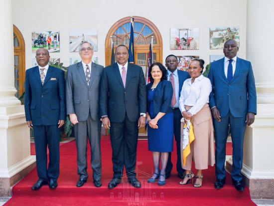 Kavi Baily is pictured with a long white shirt and long pink skirt. She stands smiling with several Kenyan officials in front of a government building.