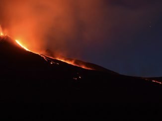 A night time scene of the top of a volcano erupting. A single stream of lava drips out the top.