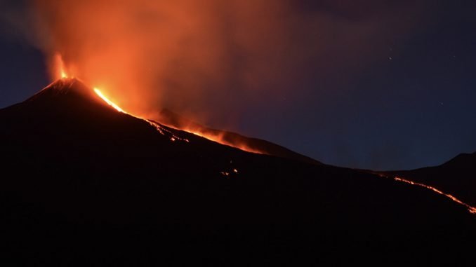A night time scene of the top of a volcano erupting. A single stream of lava drips out the top.