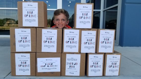 A small boy about 9 years old stands smiling behind a stack of Boxes of Love. He is blonde.