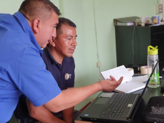 Two Latin producers look at a computer screen together. One is wearing a blue collared shirt and motioning an explanation with his hand to the producer in front of him, wearing a black collared shirt. Both are in their late 30s to early 40s.