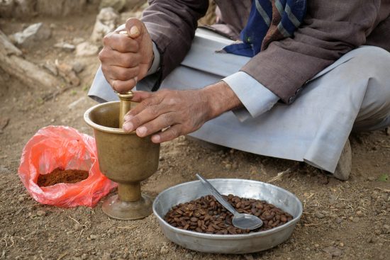 A closeup of hands churning beans in a golden mortar and pestle. A pan filled with coffee beans sits next to it.