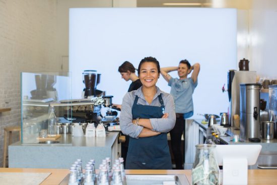 Selina is a Filipina woman in her early 40s. She poses with her arms crossed, smiling, wearing a dark blue apron and blue collared shirt. She is inside of a coffee shop with two individuals working behind her.