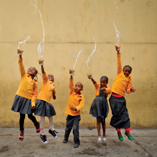 Five Ethiopian children are wearing yellow shirts and long green skirts and pants as part of their school uniforms. They throw glasses of water into the air in an artistic fashion, looking above and smiling.