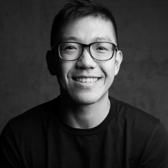 Vaughn Tan is an Asian man in his late 30s. He smiles for this black and white portrait, wearing thick frame black glasses and a black t shirt.