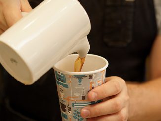 A closeup of a barista pouring coffee from a ceramic carafe into a takeaway cup.