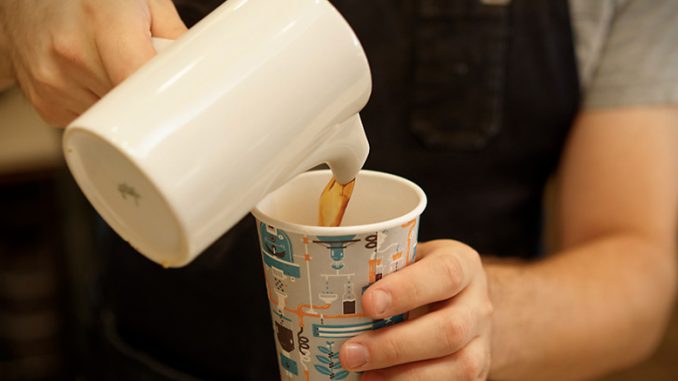 A closeup of a barista pouring coffee from a ceramic carafe into a takeaway cup.