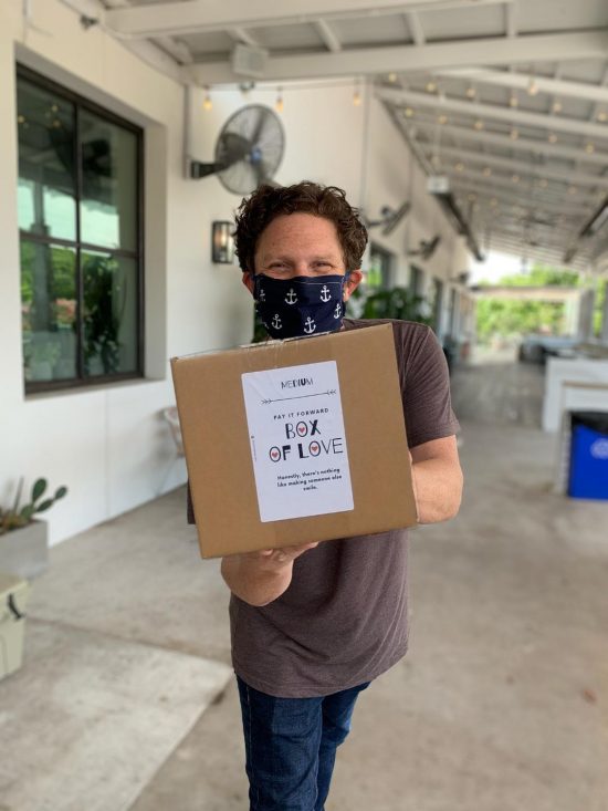 Owner Douglas Kuehn III poses with a Box of Love. He is in his 40s and has wavy short hair and wears a black face mask and brown shirt.