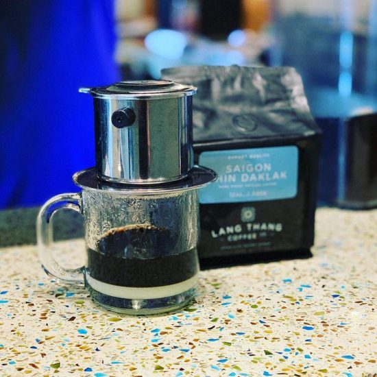 A closeup of a cafe phin Vietnamese coffee dripper brewing into a short glass mug. A bag of Vietnamese coffee from Lang Thang roasters sits next to it.
