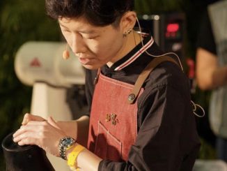 Peace is a Thai barista with short hair. She is at a coffee competition pouring water with an apron on and small mic attached.