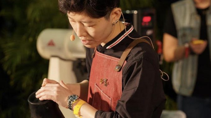 Peace is a Thai barista with short hair. She is at a coffee competition pouring water with an apron on and small mic attached.