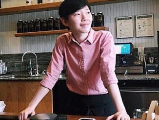 Peace is a young woman in her early 30s. She stands behind the counter of a coffee education station, wearing a pink collared shirt a smiling looking off in the distance. She has short black hair and is of Thai descent.