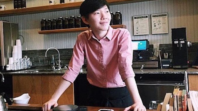 Peace is a young woman in her early 30s. She stands behind the counter of a coffee education station, wearing a pink collared shirt a smiling looking off in the distance. She has short black hair and is of Thai descent.