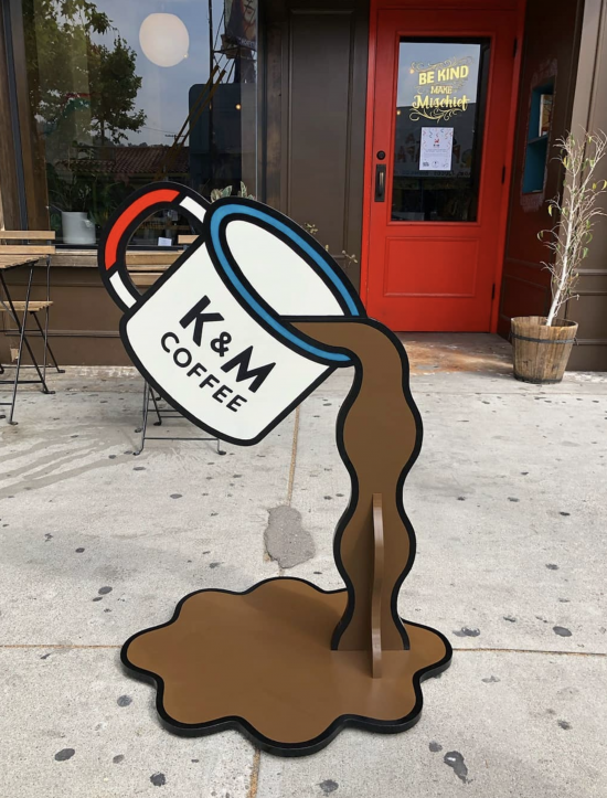 A sign outside kindness and mischief designed by remo, which features a cartoon mug spilling coffee.