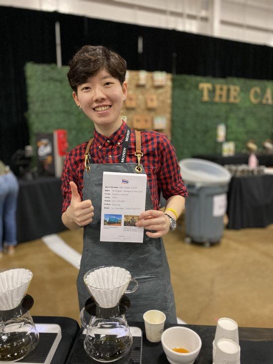 Peace poses with her coffee presentation card. She is giving a thumbs up and stands in front of pour over cones.