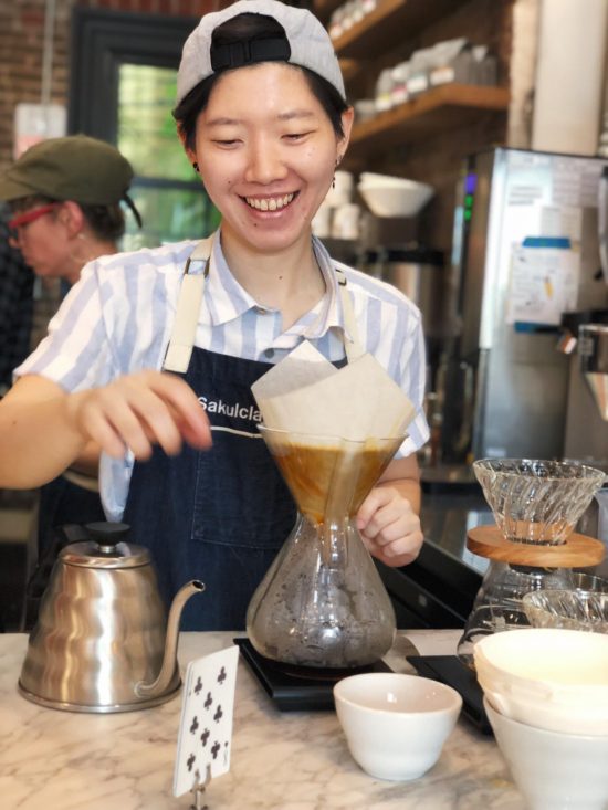 Peace smiles wearing a backwards cap. She has a blue striped collared shirt and apron, brewing a Chemex for a customer at Toby's.