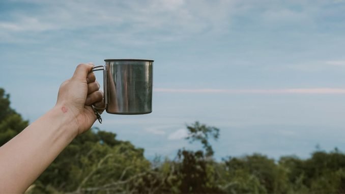 An arm holds a tin mug on top of a green mountain in Vietnam. It is a clear sky day.