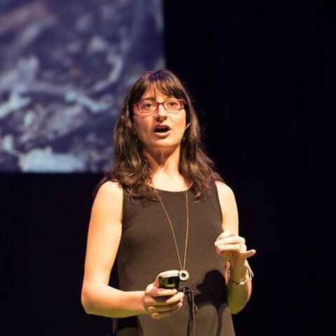 Hannah speaks at a coffee event panel on a stage. She wears glasses, and has long wavy brown hair with bangs.