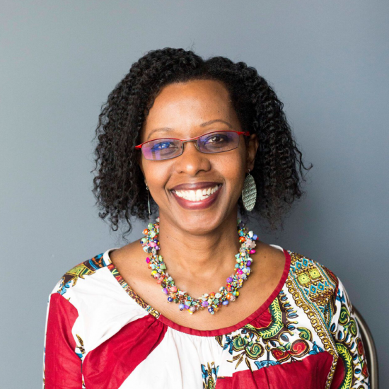 Jeanine's headshot, pictured she has tight curls, red glasses, and wears a bright red dress smiling.