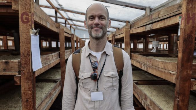 Michael Sheridan is a white male in his 40s. He smiles at the camera wearing a backpack, long sleeve collared shirt, and name tag.