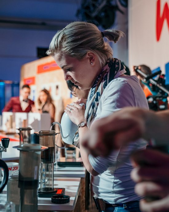 Wendelien Van Bunnik, the last AeroPress champ, stands pouring water from a kettle into the AeroPress. She is blonde and wears her hair in a pigtail and has a white t shirt on.