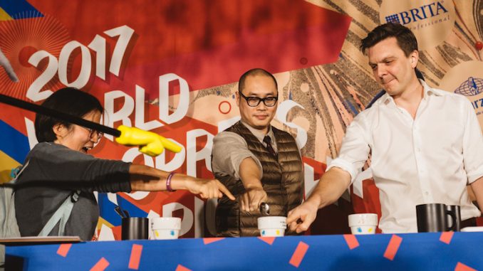 Three judges stand in a circle pointing at a cup of AeroPress coffee.