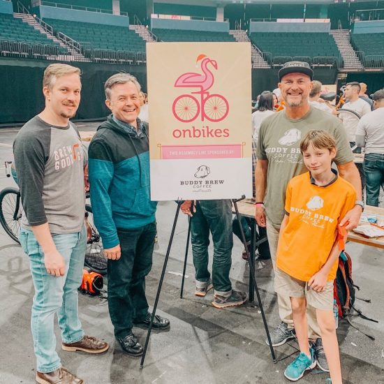 A group of people stand smiling in a stadium in front of an Onbikes sign at a bike build event.