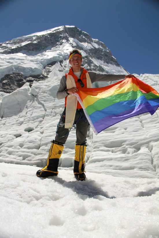 Cason is a young 19 year old in this photo, blonde and smiling on top of a mountain. He proudly holds the rainbow Pride flag.