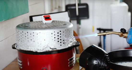 A blow torch blows flames onto a rice cooker with a steam top basket on it wrapped in foil.