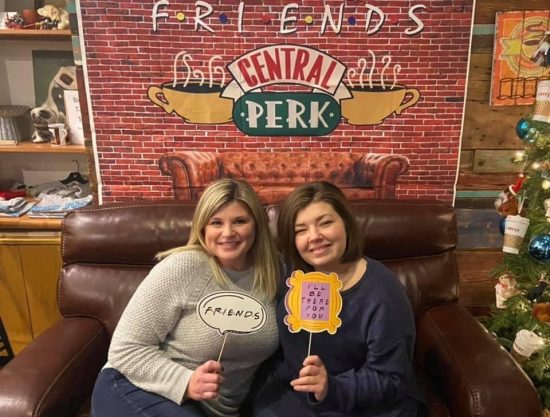 Two people, one blonde and one brunette, stand smiling in front of a sign that refers to the central perk store from friends.
