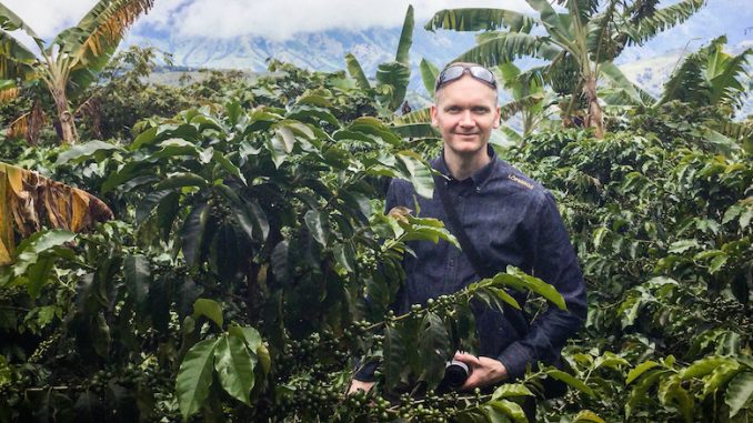 A man stands smiling amidst a sunny day in a jungle full of plants.