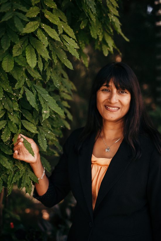 Dr. Sarada Krishnan is a woman in her early 40s. She has bangs and long hair and smiles next to a coffee plant.