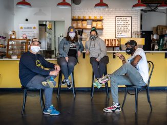 Four members who work at Enderly smile sitting on chairs spread out at the front of Enderly Coffee's bar. They are wearing face masks.