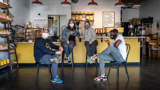 Four members who work at Enderly smile sitting on chairs spread out at the front of Enderly Coffee's bar. They are wearing face masks.