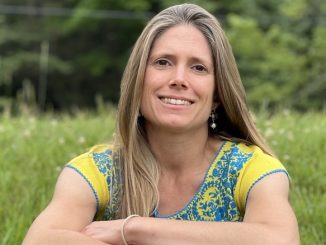 Alyson Welch in a blonde white woman in her 40s. She poses with her arms crossed smiling in a grassy field.