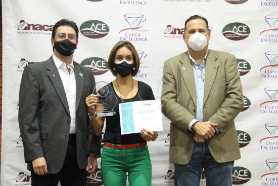 Three people stand in front of a cup of excellence banner wearing face masks and holding their certificate that they won.