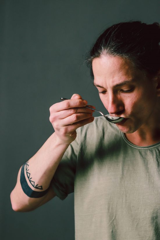 Alan in a portrait image, closing his eyes and sipping on a spoon filled with coffee. His hair is tied back in a pony tail.