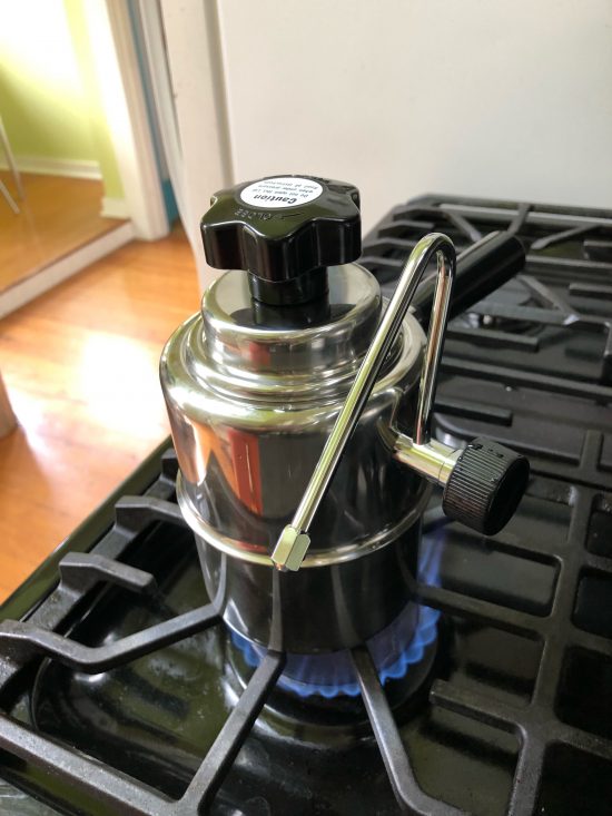 The stovetop steamer, a short cylindrical body with a 3 inch handle and steam wand attached sits on top of a black and silver stove.