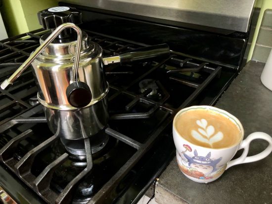 The cup of coffee with milk poured into it sitting next to the stovetop steamer in a kitchen.