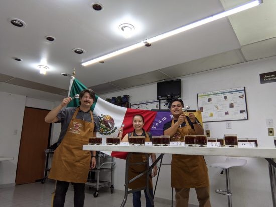 Cup of Excellence winners stand smiling on a screen on video. The flag of Mexico is behind them.