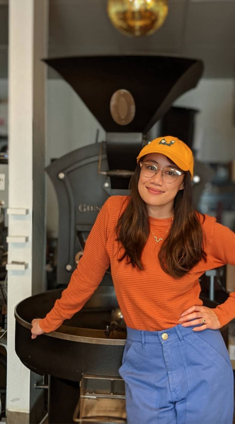 Mo stands in front of a coffee roaster. She wears a bright orange baseball cap and and orange sweater.