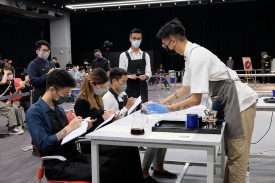 Three judges sit with face masks on writing on clipboards. A competitor is presenting their coffee to the judges.