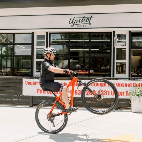 A man wears a bike helmet and holds his bike upright with him on it in front of a coffee shop with big windows.