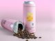Two cans next to each other, one standing and the other spilled with coffee beans coming out of it. It is against a pink background.