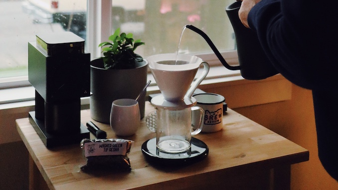 A hand pouring water from a goose neck kettle over a sleek pour over cone on a home coffee station.