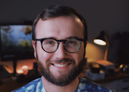 Caleb Stultz is a white man in his late 20s. He smiles for a portait. He has thick framed glasses, brunette hair, and a thick brunette beard.