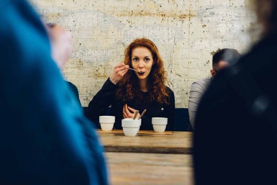 A woman with red hair sits at a table with a cupping spoon held up to her mouth. There are three white cupping bowls in front of her.